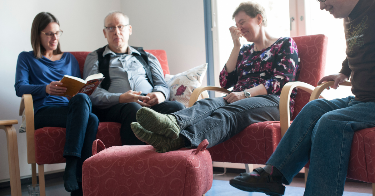En person läser högt för tre andra personer. De sitter i mjuka fåtöljer och ser glada ut.